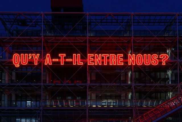 la façade du Centre Pompidou à Paris portant une immense enseigne de néon rouge avec la question en lettres capitales : "QU'Y A-T-IL ENTRE NOUS ?"